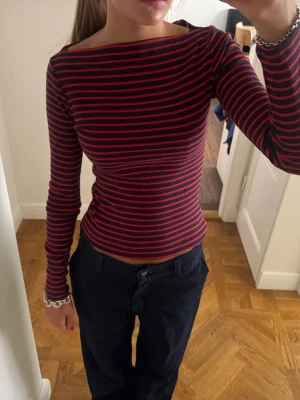 Randig långärmad topp i rött och blått - Säljer en slim långärmad topp med tunna röda och marinblå ränder. Toppen har en båtringad hals och sitter tight mot kroppen. Perfekt att styla med jeans eller kjol för en cool vardagslook.