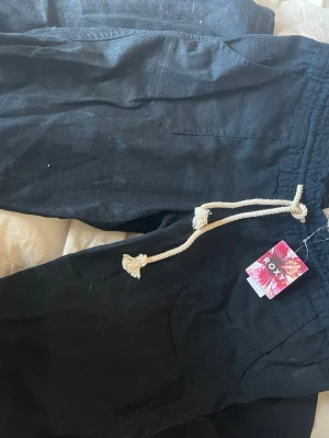 Svarta linnebyxor från Roxy - Snygga svarta linnebyxor från Roxy med avslappnad passform och elastisk midja med vit dragsko i rep. Byxorna har två fickor fram och två bak, samt en liten broderad logga på ena bakfickan. Perfekta för en chill och stilren look.