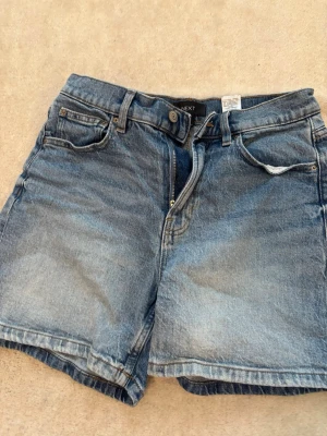 Blå jeansshorts från NEXT - Klassiska blå jeansshorts från NEXT med fem fickor och knappgylf. Shortsen har en avslappnad passform och är tillverkade i bomull med en snygg tvättad look. Perfekta för varma dagar och enkel att matcha med olika stilar.