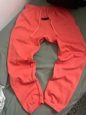 Korallrosa Essentials mjukisbyxor - Snygga korallrosa mjukisbyxor från Essentials med broderad logga på benet. Byxorna har resår i midjan med dragsnöre och elastiska benslut. Perfekta för chill och streetwear, tillverkade i mjukt bomullsmaterial.