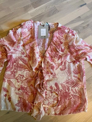 Blommig blus från Design by Si - Blus i tunn, luftig kvalitet från Design by Si. Mönstrad i rosa och vitt med stora blommor och blad. Blusen har knytband framtill, v-ringning och trekvartsärm. Perfekt för sommaren.