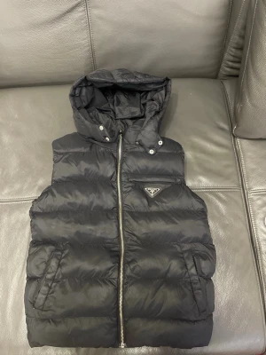 Prada väst  8-9 134-160 - Snygg svart dunväst från Prada med avtagbar huva och dragkedja framtill. Västen har två fickor och den ikoniska Prada-loggan på bröstet. Perfekt för lager-på-lager och streetstyle. Tillverkad i glansigt material som ger en exklusiv känsla. Storlek 8-9 134-160