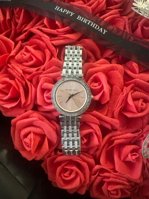 Michael Kors silverfärgad klocka - Stilren klocka från Michael Kors med silverfärgat armband i metall och rund urtavla i ljusrosa. Klockan har gnistrande stenar runt boetten för extra bling och ett klassiskt, elegant utseende. Perfekt accessoar för att lyfta din outfit. Några stenar saknas 