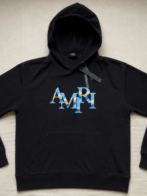 Svart hoodie från Amiri med logga - Snygg svart hoodie från Amiri med stor färgglad AMIRI-logga i blått och gult på bröstet. Klassisk känguruficka framtill och huva. Tillverkad i mjukt material som känns skönt mot huden. Perfekt för dig som gillar streetwear och vill sticka ut.