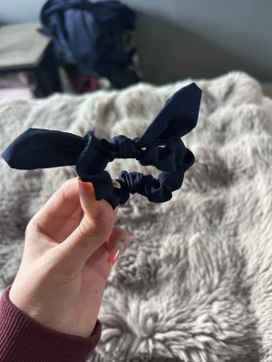 Mörkblå scrunchie med rosett - Säljer en mörkblå scrunchie med söt rosettdetalj. Hårsnodden är gjord i ett mjukt tyg som ger en snygg look och håller håret på plats. Perfekt accessoar för att piffa upp din frisyr.