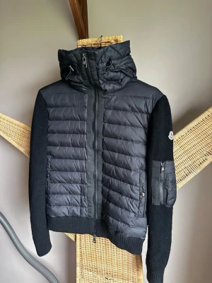 Moncler Knitted Jacket Size 1 - cardiganjacka från Moncler med vadderad framsida och huva i nylon samt stickade ärmar och rygg. Jackan har dragkedja framtill, fickor med dragkedja och Moncler-logga på ärmen. Kvitto saknas.
