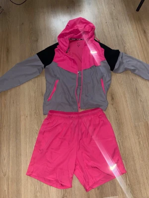 Nike träningsset jacka och shorts rosa - Nike träningsset med en grå och rosa vindjacka med huva och dragkedja samt matchande rosa träningsshorts. Jackan har svarta detaljer på axlarna och fickor med dragkedja. Perfekt för löpning eller träning utomhus.