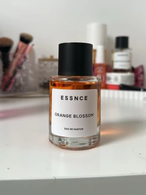 ESSNCE Orange Blossom parfym - Fräsch parfym från ESSNCE med doft av orange blossom. Flaskan är cylindrisk i genomskinligt glas med svart lock och vit etikett. Innehållet har en varm, bärnstensfärgad ton. Perfekt för dig som gillar blommiga och fruktiga dofter.