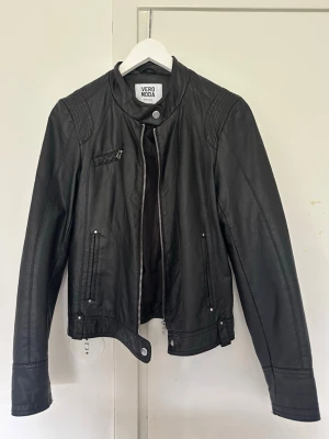 Svart skinnjacka från Vero Moda - Snygg svart skinnjacka från Vero Moda med ståkrage, dragkedja framtill och coola detaljer på axlarna. Jackan har en liten bröstficka. Perfekt för en cool look och passar till det mesta.