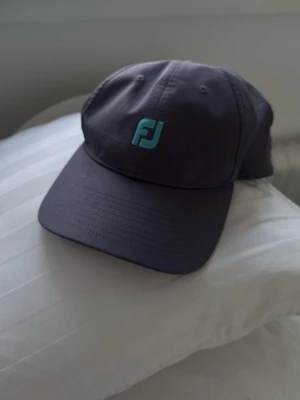 Keps från FootJoy med logga - Snygg keps från FootJoy med turkos logga framtill och justerbart band bak med blå och vita ränder. Kepsen har klassisk böjd skärm och är tillverkad i ett lätt och bekvämt material, perfekt för en sportig look. 