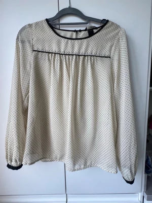 Prickig blus från H&M - Söt vit blus från H&M med små svarta prickar och svarta detaljer vid halsringning och ärmslut. Långärmad modell med lätt transparent tyg. 