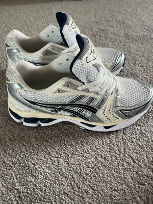 Vita och silver Asics sneakers - Snygga sneakers från Asics i vitt och silver med Blå detaljer. Endast använda en gång för en promenad med hunden, skorna är i storlek 42 och är true too size. Använder inte då det är för små annars tvär feta.