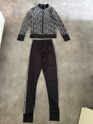 2-delatset. Ziptröja med byxor - 2-delat set. Ziptröja med bokstäver på. Svarta byxor/leggings med text på sidan. Hela setet är köpt i S. Aldrig använt!