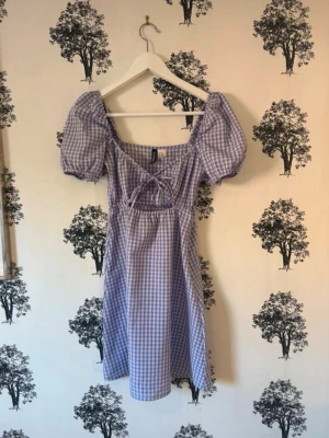 Lila rutig klänning med puffärmar - Superfin lila och vit rutig klänning med puffiga kortärmar och söt knytning framtill. Klänningen har en fyrkantig urringning och är i en lätt, luftig bomullskvalitet. Perfekt för vår och sommar!