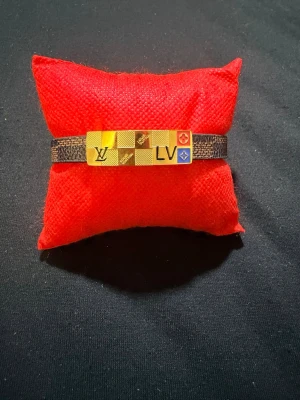 Rostfritt stål armband med gulddetaljer - Snyggt armband i brunt läder med guldfärgade i rostfritt stål. Armbandet har rutmönstrade detaljer och färgade symboler på framsidan samt ett elegant spänne i guld på baksidan. Perfekt accessoar för dig som gillar exklusiv stil.