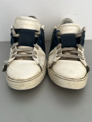 Valentino Garavani vita och blå sneakers - Snygga sneakers från Valentino Garavani i vitt och marinblått läder. Skorna har klassisk låg profil, vita snören och tydlig logga på plösen. Perfekta för dig som gillar exklusiva och stilrena detaljer.