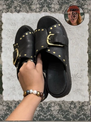 Svarta sandaler med gulddetaljer - Säljer ett par svarta sandaler i läder-look med breda remmar, stora guldfärgade spännen och nitar. Snygg chunky sula och öppen tå. Perfekta för dig som gillar en edgy och trendig stil.