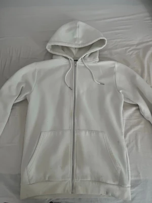 Vit zip hoodie smog - Säljer en enkel och clean vit hoodie med dragkedja framtill och två stora fickor. Hoodien har justerbar dragsko i huvan och ribbade muddar vid ärmar och nederkant. Perfekt för dig som gillar en stilren look. Materialet känns mjukt och bekvämt. Har inte tvättat tröjan ännu så ser lite risig ut på bild kanske
