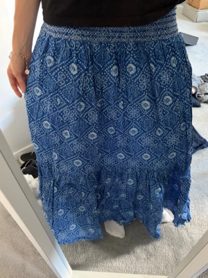 Blå långkjol med mönster - Säljer en blå långkjol med vitt paisley-inspirerat mönster. Kjolen har bred, smockad midja och faller i mjuka volanger. Perfekt för dig som gillar bohemisk stil och vill ha något bekvämt och snyggt till sommaren. Passar mig som har S/M. 