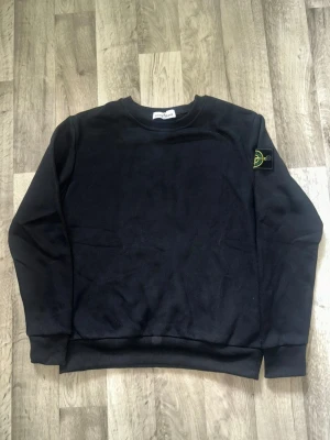 Svart sweatshirt från Stone Island - En svart sweatshirt från Stone Island med klassisk rund hals och ribbade muddar. Tröjan har den ikoniska Stone Island-loggan på vänster ärm. Perfekt för en clean och stilren look.
