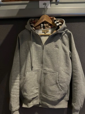 Grå zip hoodie från Burberry - Snygg grå zip hoodie från Burberry med klassiskt rutigt foder i huvan och broderad logga på bröstet. Hoodien har dragsko i huvan, två stora fickor fram och ribbade muddar. Perfekt för en chill och stilren look.