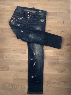 Mörkblå jeans med blommor - Snygga mörkblå jeans med rosa broderade blommor och slitna detaljer. Jeansen har knappgylf, fem fickor och vita färgstänk för en trendig look. Perfekta för dig som vill ha något unikt i garderoben.