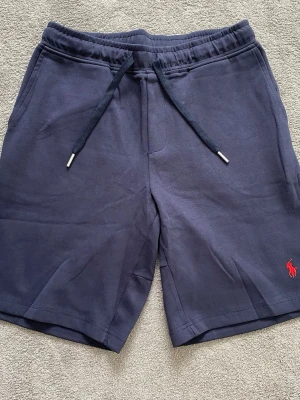 Marinblå shorts från Polo Ralph Lauren - Snygga marinblå shorts från Polo Ralph Lauren med klassisk röd logga broderad på benet. Tillverkade i mjuk bomull för maximal komfort. Perfekta för en avslappnad och stilren look. Storlek M. 