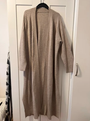 Beige lång stickad kofta - Lång, ribbstickad kofta i beige med öppen front och långa ärmar. Perfekt för lager på lager och ger en chill vibe till din outfit. Mjuk och skön, passar till det mesta i garderoben.