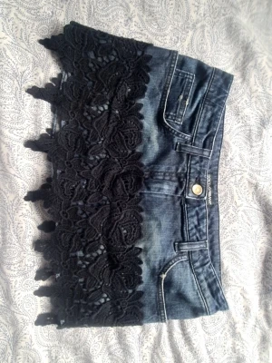 Mini Jeanskjol med spets från Dolce & Gabbana - Unik kort jeanskjol från Dolce & Gabbana med mörkblå denim och bred svart spetsvolang nedtill. Kjolen har klassiska jeansdetaljer som fickor och knapp, och spetsen ger en extra stylish touch. På bild 5 ser man små vita trådar som sticker ut, samt på näst sista bilden ser man nått slitage på framfickan, som också finns liknande på andra framfickan. 