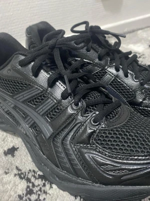Asics Gel Kayano 14 - Säljer ett par helsvarta Asics sneakers med klassisk logga på sidan. Skorna har en kombination av mesh och syntetläder, vilket ger både snygg look och bra ventilation. Tjock sula och snörning för skön passform. Perfekta för dig som gillar sportig stil.