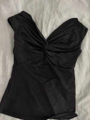 Svart baddräkt med twistad front - Stilren svart baddräkt med twistad detalj framtill och breda axelband. Klassisk modell med mjukt, stretchigt material som ger en smickrande passform. Perfekt för strand eller poolhäng.