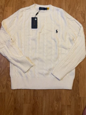 Vit kabelstickad tröja Polo Ralph Lauren - Kabelstickad vit tröja från Polo Ralph Lauren med klassisk rund halsringning och broderad marinblå logga på bröstet. Tröjan har ribbade muddar vid ärmslut och nederkant, och är tillverkad i mjukt material som passar perfekt till lager på lager.