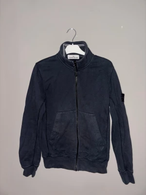 Svart ziptröja från Stone Island - Snygg svart ziptröja från Stone Island med hög krage och ikonisk patch på vänster ärm. Tröjan har dragkedja framtill, ribbade muddar och är tillverkad i mjukt bomullsmaterial. Perfekt för en streetwear-look.