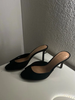 Svarta öppna pumps från Mango - Stilrena svarta pumps från Mango med öppen tå och smal klack. Skorna har en klassisk slip-in modell och är tillverkade i mockaimitation med beige innersula. Perfekta för dig som gillar en elegant och enkel look.