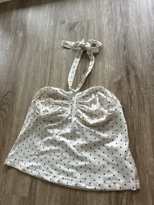 Vit prickig halterneck linnetopp - Supersöt vit linnetopp med svarta prickar och halterneck med knyt i nacken. Toppen har rynkade kupor och ärmlös design, perfekt för varma dagar. Mjuk och stretchig bomullsblandning som sitter bekvämt.