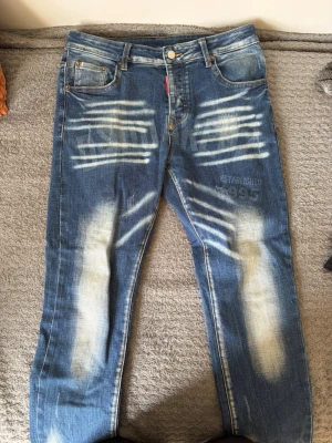 Blå distressed jeans från Dsquared2 - Säljer ett par blå jeans från Dsquared2 med kraftiga slitningar och blekta partier framtill och baktill. Jeansen har tryck med texten 'ESTABLISHED 1995' och Dsquared2-loggor, klassisk femficksmodell och raka ben. Materialet är denim i bomull.