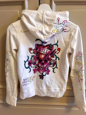 White zip hoodie Don Ed Hardy with skull design  - Super cool Ed Hardy zip hoodie in white with a pink skull design decorated with sparkling rhinestones. Iconic graphics on the front, back, and sleeves. Zip-up style with a hood, soft and comfortable to wear. (The size runs small!)                It’s a size M, but I think it runs small and fits more like an XS–S.