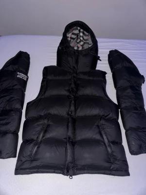 Svart dunväst från Burberry - Snygg svart dunväst från Burberry med avtagbara ärmar och huva. Västen har dragkedjor på fickorna och klassiskt rutigt foder i huvan. Perfekt för lager på lager och streetwear-vibe.