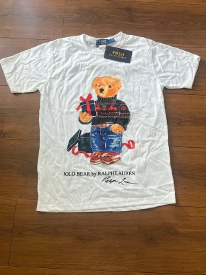 Vit Polo Bear t-shirt från Ralph Lauren - Vit t-shirt från Ralph Lauren med ikoniskt Polo Bear-motiv framtill. Björnen har på sig en mönstrad tröja, jeans och håller i ett presentpaket. T-shirten har rund hals och korta ärmar. Perfekt för dig som gillar klassiska och lekfulla prints.