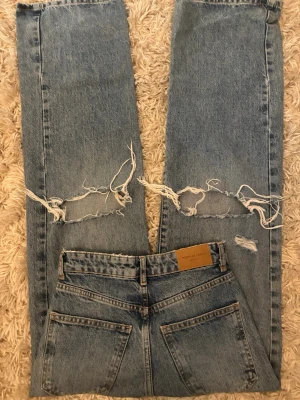 Blå jeans med hål och raka ben - Säljer ett par blå gina jeans med raka ben och stora slitningar över knäna. Klassisk femficksmodell i kraftig denim med hög midja och råa kanter vid hålen. Perfekta för en avslappnad och trendig look.
