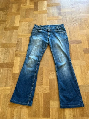 Blå bootcut jeans med slitningar - Säljer ett par blå bootcut jeans med snygga slitningar framtill och klassiska bakfickor. Jeansen har låg midja och är tillverkade i denim med en lätt tvättad look. Perfekta för en avslappnad och trendig stil.