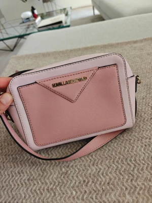 Rosa crossbody från Karl Lagerfeld - Snygg rosa crossbody-väska från Karl Lagerfeld med justerbar axelrem och guldfärgade detaljer. Väskan har en stilren design med en ficka framtill och logga i metall. Tillverkad i slittåligt konstläder med två olika rosa nyanser.