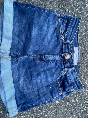 Blå denimshorts från Missguided - Säljer ett par klassiska blå denimshorts från Missguided med hög midja och uppvikta benslut. Shortsen har fem fickor, bälteshällor och stängs med knapp och dragkedja. Perfekta för sommaren och enkla att matcha med olika stilar.