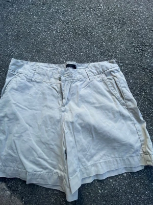 Beiga shorts från Gina Tricot - Säljer ett par beiga shorts från Gina Tricot med klassisk passform och fickor på sidorna. De har bälteshällor och stängs med dragkedja och knapp. Perfekta för varma dagar och enkla att matcha med olika toppar.