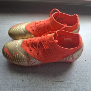 Puma fotbollsskor röd/guld - Snygga fotbollsskor från Puma i en cool kombination av rött och guld. Skorna har en strumpliknande passform med stickad ovandel och snörning för bra stöd. Yttermaterialet är syntetiskt och skon har Pumas logga på sidan. Perfekta för dig som vill sticka ut på planen.