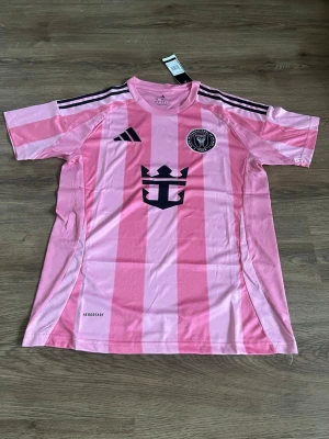 Rosa Inter Miami CF matchtröja Adidas - Säljer en rosa Inter Miami CF matchtröja från Adidas med svarta detaljer och klubbmärke på bröstet. Tröjan har svarta ränder på axlarna, korta ärmar och är tillverkad i lätt, ventilerande material med Aeroready-teknologi. Perfekt för fotboll eller träning.