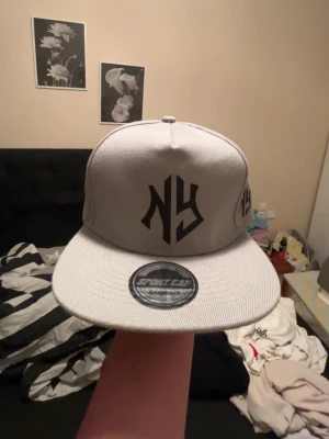 Grå snapback keps med NY-logga - Snygg grå snapback keps med svart NY-logga framtill och på sidan. Kepsen har platt skärm och justerbar baksida. Tillverkad i ett slitstarkt tyg som passar perfekt till streetwear-stilen.