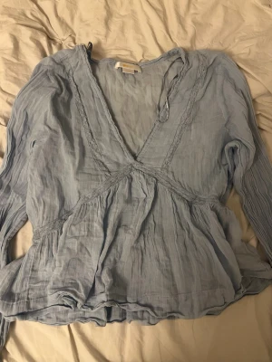 Ljusblå blus från Gina Tricot - Supersöt ljusblå blus från Gina Tricot med v-ringning och tunna spetsdetaljer. Blusen har lång ärm och är gjord i ett lätt, luftigt tyg som ger en avslappnad och flowy känsla. Perfekt att styla med jeans eller kjol för en trendig look.