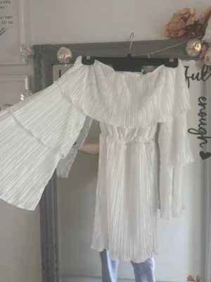 Vit offshoulder klänning med volang - Superfin vit offshoulder klänning med bred volang runt axlarna och långa, vida ärmar med volangdetaljer. Klänningen har plisserat tyg och resår i midjan för en snygg passform. Perfekt för sommarens alla tillfällen!