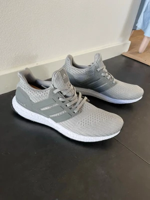 Grå Adidas Ultraboost sneakers - Snygga grå Adidas Ultraboost sneakers med stickad ovandel i mesh för bra andningsförmåga. Skorna har klassiska tre ränder på sidan, vit Boost-sula för extra komfort och svart yttersula. Perfekta för dig som vill ha både stil och funktion.